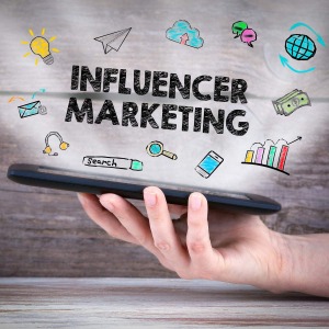 Influencer Marketing auf einem Bild dargestellt. Es wird ein Handy in der Hand gehalten und darüber schweben sämtliche Zeichen die auf Social Media hinweisen.